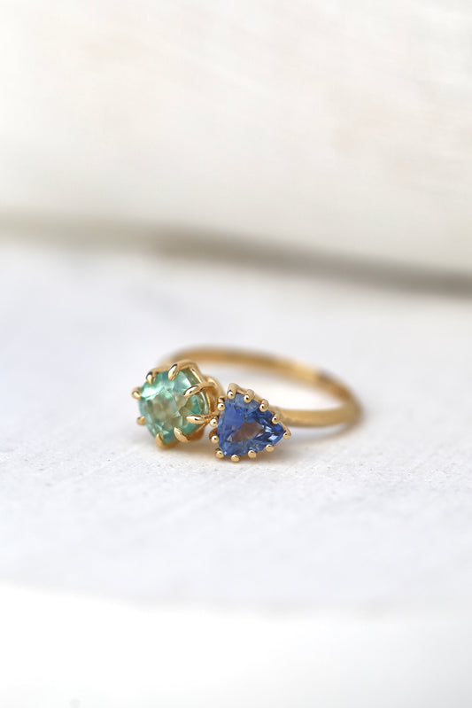 18K Toi et Moi ring features a cushion-cut green tourmaline paired with a triangular blue sapphire, creating a gorgeous and balanced contrast. Each stone is set in a distinct style: the green tourmaline in a claw prong setting, and the sapphire in a classic round prong setting.