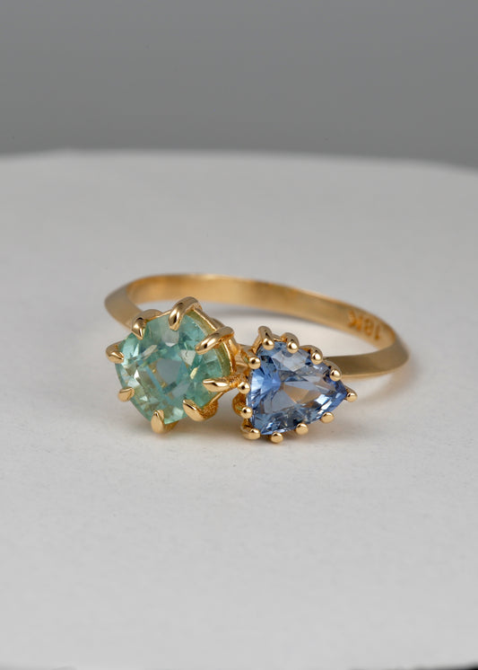 18K Toi et Moi ring features a cushion-cut green tourmaline paired with a triangular blue sapphire, creating a gorgeous and balanced contrast. Each stone is set in a distinct style: the green tourmaline in a claw prong setting, and the sapphire in a classic round prong setting.