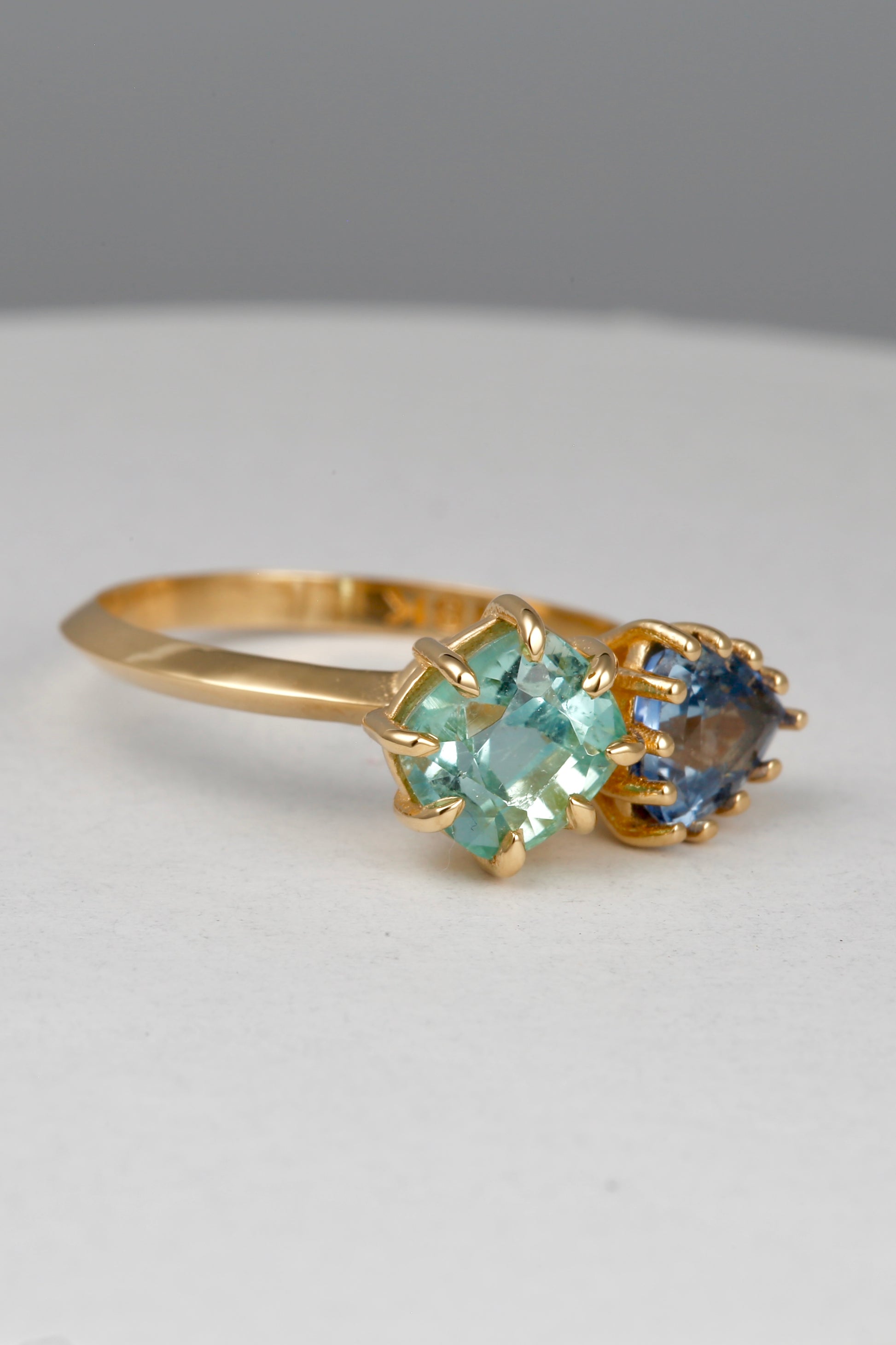 18K Toi et Moi ring features a cushion-cut green tourmaline paired with a triangular blue sapphire, creating a gorgeous and balanced contrast. Each stone is set in a distinct style: the green tourmaline in a claw prong setting, and the sapphire in a classic round prong setting.