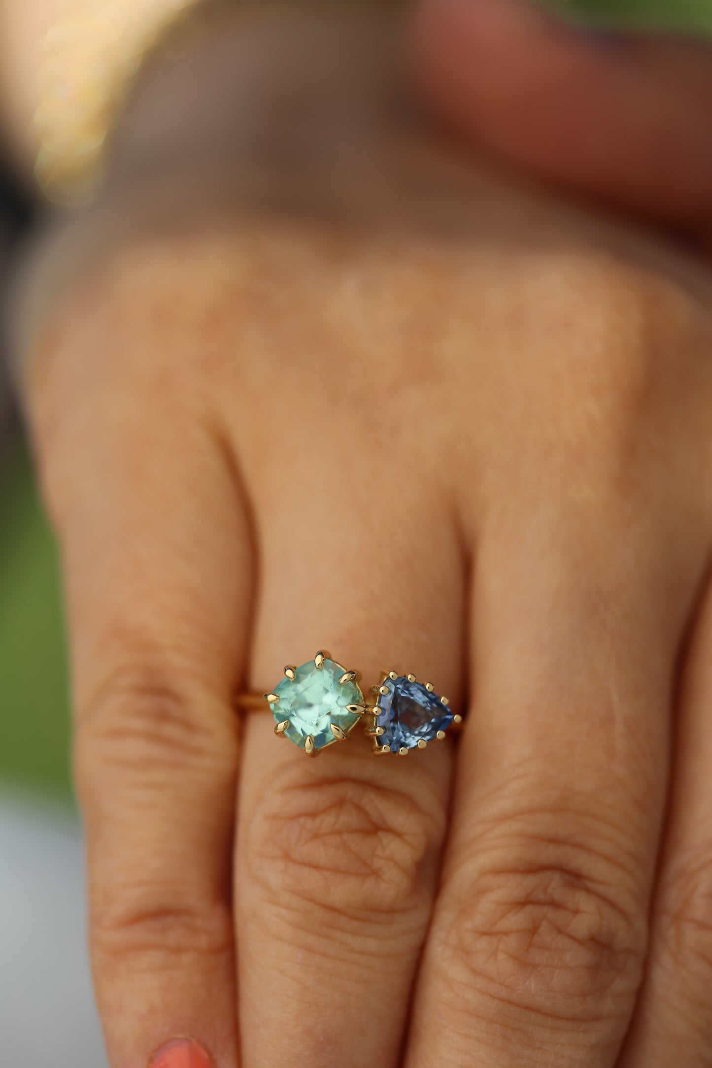 18K Toi et Moi ring features a cushion-cut green tourmaline paired with a triangular blue sapphire, creating a gorgeous and balanced contrast. Each stone is set in a distinct style: the green tourmaline in a claw prong setting, and the sapphire in a classic round prong setting.
