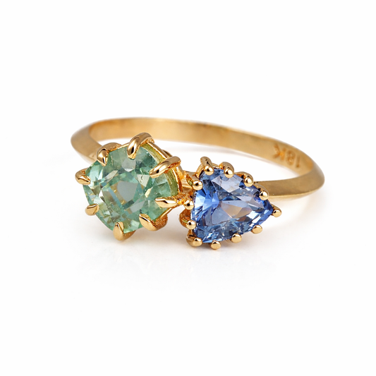 18K Toi et Moi ring features a cushion-cut green tourmaline paired with a triangular blue sapphire, creating a gorgeous and balanced contrast. Each stone is set in a distinct style: the green tourmaline in a claw prong setting, and the sapphire in a classic round prong setting.