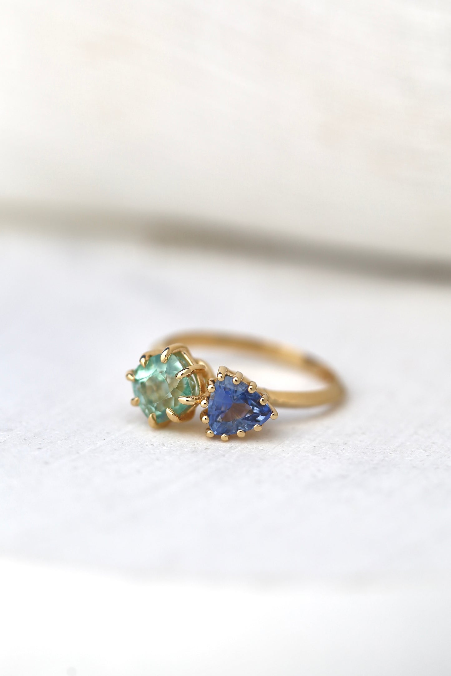 18K Toi et Moi ring features a cushion-cut green tourmaline paired with a triangular blue sapphire, creating a gorgeous and balanced contrast. Each stone is set in a distinct style: the green tourmaline in a claw prong setting, and the sapphire in a classic round prong setting.