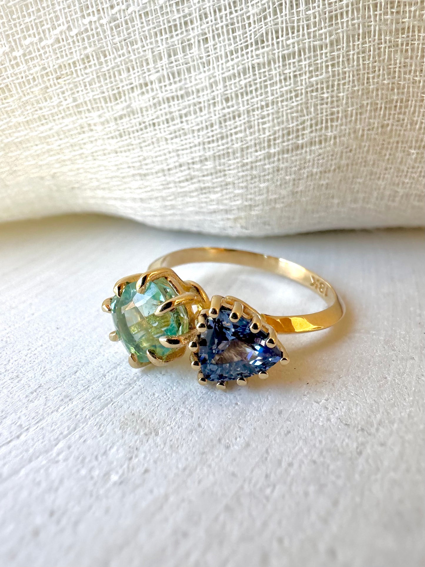 18K Toi et Moi ring features a cushion-cut green tourmaline paired with a triangular blue sapphire, creating a gorgeous and balanced contrast. Each stone is set in a distinct style: the green tourmaline in a claw prong setting, and the sapphire in a classic round prong setting.