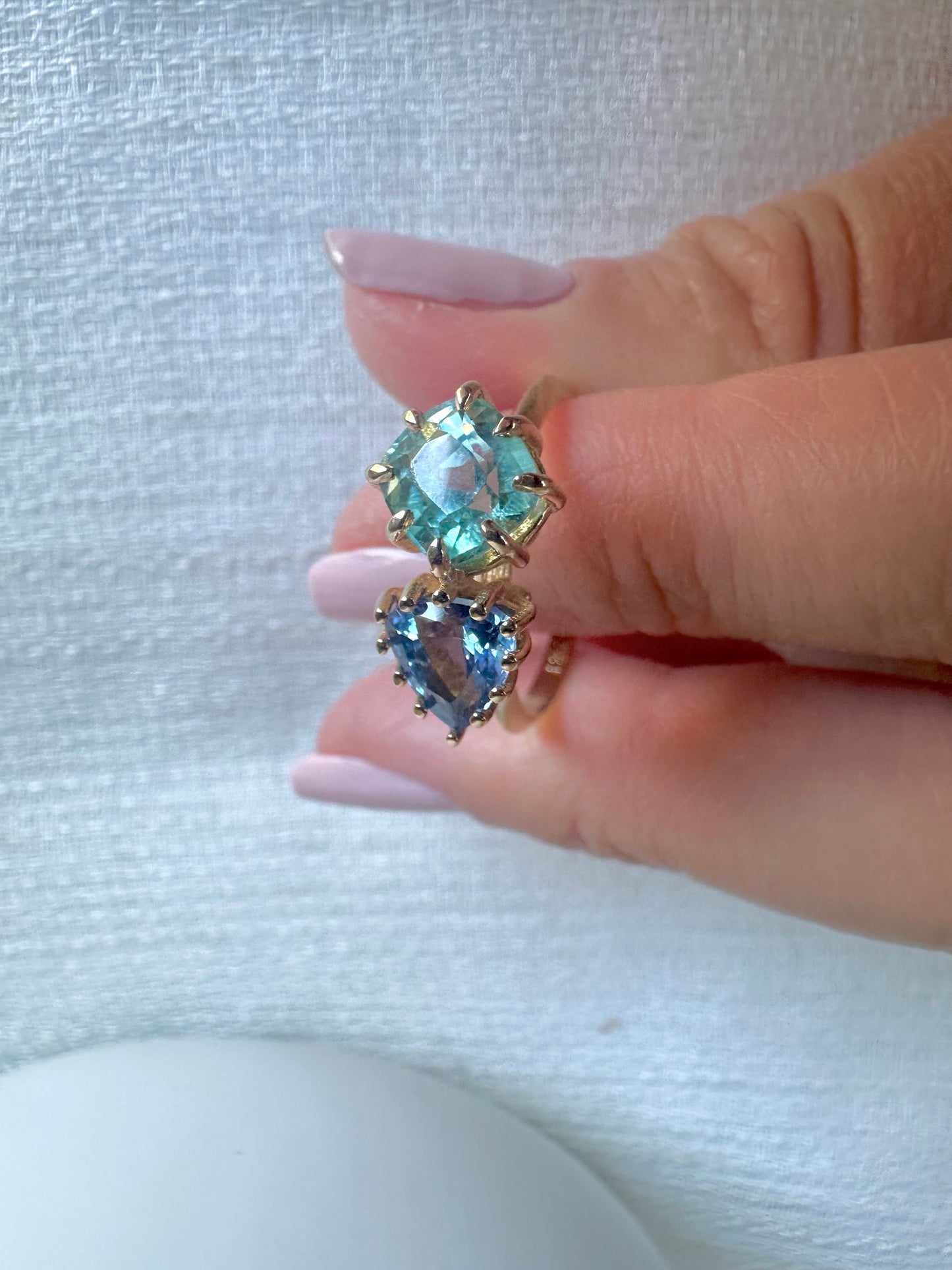 18K Toi et Moi ring features a cushion-cut green tourmaline paired with a triangular blue sapphire, creating a gorgeous and balanced contrast. Each stone is set in a distinct style: the green tourmaline in a claw prong setting, and the sapphire in a classic round prong setting.